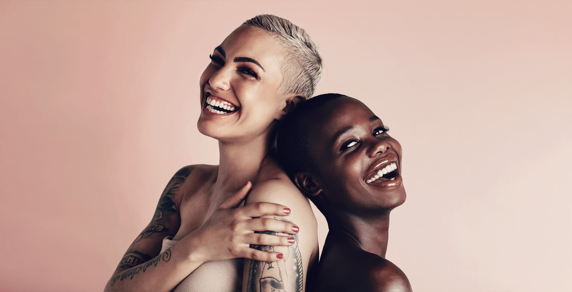 Two women smiling with clean beauty products on their faces on a peach background.