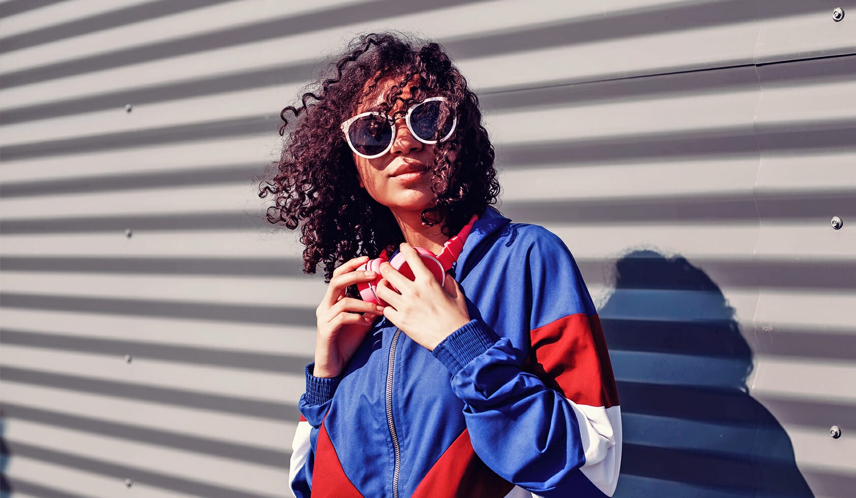 Woman posing with headphones and a 90s themed Y2K jacket against a metal wall.